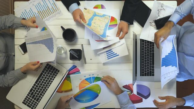 Top view to hands of business people sitting at table in trendy office and examining graphs. Creative team working efficiently on business development. View of working mess on office desk. Slow motion