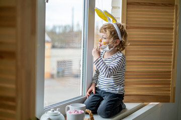 Easter 2021 concept: A child in home quarantine plays by the window with the ears of a rabbit on his head wearing a medical mask against viruses during the coronavirus COVID-2019 and the flu outbreak.