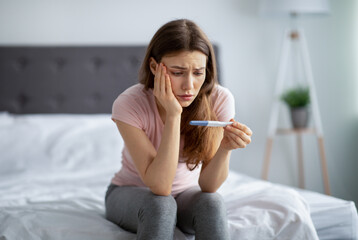 Sad young woman checking her recent pregnancy test, sitting on bed at home