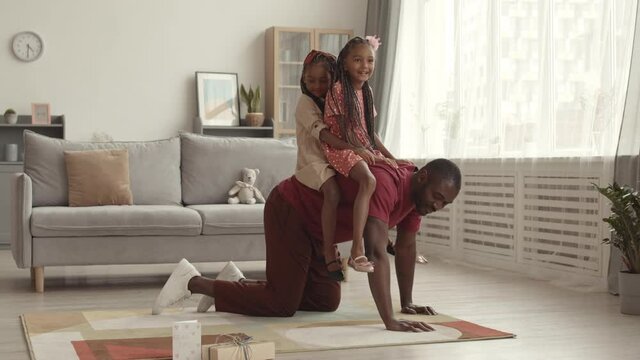 Wide shot of young African father walking on all fours in living room, riding two little adorable female kids on his back, smiling