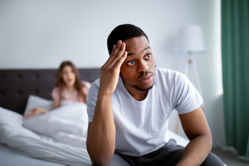 Disappointed African American guy sitting on bed, listening to his wife's quarreling, holding his...
