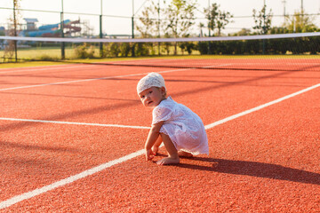 One-year-old sweet girl in white dress walks on red tennis court in summer