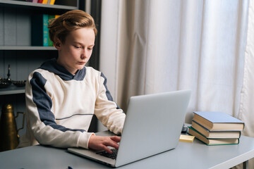 Close-up of focused pupil boy using typing laptop computer doing online lesson via Internet sitting at desk near window, side view. Concept of distance education at home