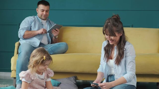 Little girl playing with mother on the carpet, while her father works using a tablet. Spending time together at home. Mom is always there to help.