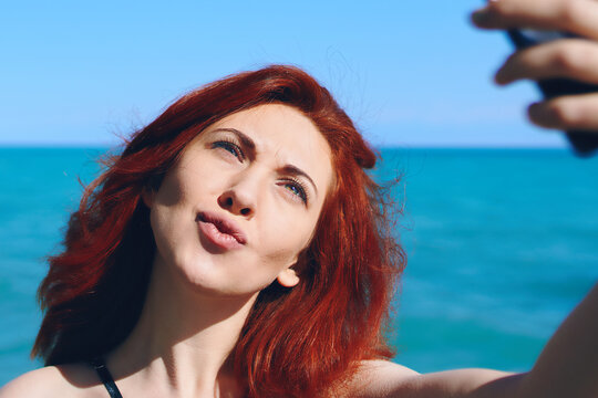 Red-haired woman makes her lips duck and takes selfie on smartphone camera. Summer sunny vacation. Pretty female takes pictures of herself against background of sea and sky.