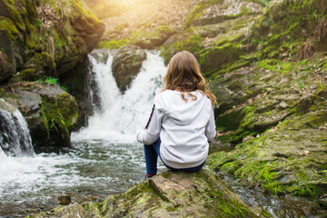 Young girl sitting on the rock and looking at waterfall on the river in the rocks. Tourism with...