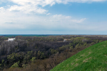 Obraz premium A beautiful spring landscape with a green forest and green meadow grass in the foreground against the backdrop of a winding river between fields and hills and a blue, slightly cloudy sky.
