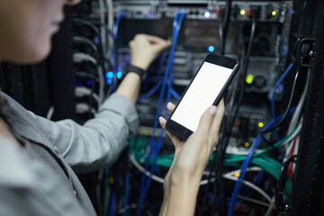 Close up of female network technician holding smartphone with blank screen while connecting cables in server cabinet, copy space