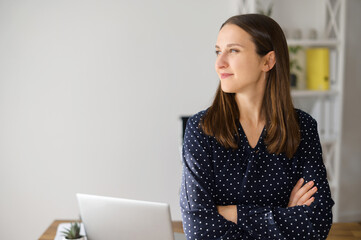 Positive female entrepreneur stands with arms crossed in modern office, confident woman in casual...