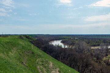 Obraz premium Panoramic view from the high bank to the river, forest, green meadow and beautiful fields. View from the high bank to the spring river and the trees around.