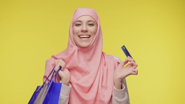 Smilling young Muslim woman in hijab headscarf posing look at camera hold in hand credit bank card and hold in hands package bag with purchases after shopping isolated on yellow background in studio.
