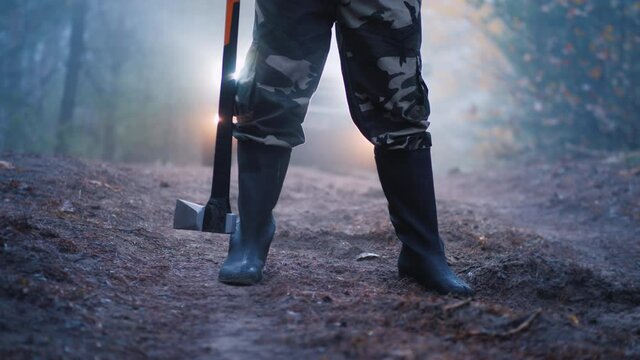 Legs in boots of a man who holds an ax, car headlights shine from behind