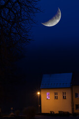Duży księżyc na nocnym niebie nad domem, w którym pali się fioletowe światło.