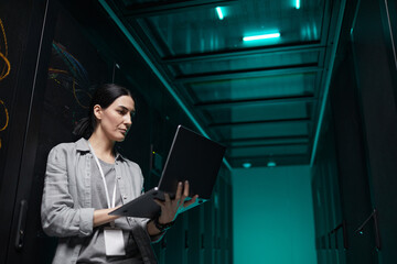 Waist up portrait of female data engineer holding laptop while working with supercomputer in server...