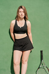 beautiful young red-haired girl dressed in a black skirt and top on a tennis court with her racquet next to her and a green wall in the background