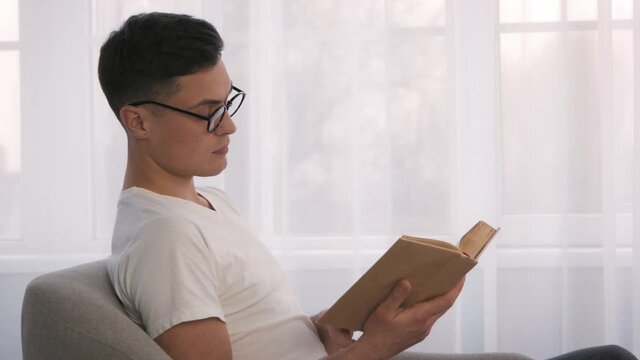 Adult bookworm. Young focused man nerd in eyeglasses reading book at home, sitting in armchair, side view, slow motion
