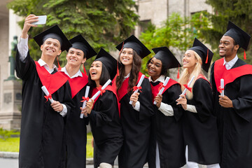 Happy graduates taking selfie together at university campus, using smartphone