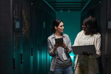 Waist up portrait of two young women using laptop while working with supercomputer in server room,...