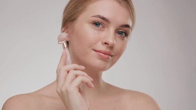 Close up portrait smiling young woman using jade facial roller for skin care, beauty treatment on white isolate background. Blonde girl using natural massager. Face massage.