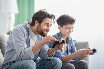 Common hobby. Overjoyed father and son playing video games and competing with each other, sitting on sofa