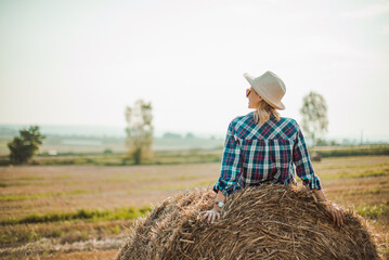 Naklejka na ściany i meble Woman in hat sitting on round bales in the field, enjoying good weather. Beautiful landscape for young entrepreneurs farmers. Back view image in the summer.