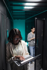 Vertical portrait of female data engineer using laptop in server room while setting up...