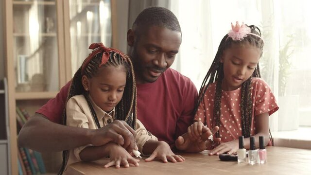 Medium shot of young happy African man sitting at table, watching his two little cute daughters playing with makeup, painting his nails with polish