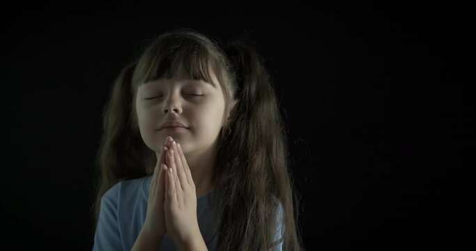 The kid is praying. Beautiful little girl prays on a black background.