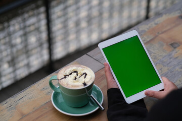 over shoulder view of hand holding green screen tablet. Cup of coffee on cafe table 
