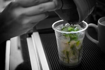 Barista's hand pouring matcha into a glass of ice cube and milk. Close up of making matcha latte for customer at coffee shop. Black and white tone except a glass of matcha latte