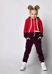 little blonde curly-haired beautiful girl in a red burgundy tracksuit and white sneakers stands with her hands in her pockets and is shy. over gray wall background. Stylish casual fashion for kids.