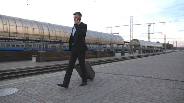 Handsome businessman walking through railway station with his luggage and talking on phone.Young business person strolling near railway and having work conversation. Concept of business trip. Slow mo