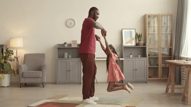 Full shot of young strong African man standing in room, circling his seven-year-old daughter around, holding by arms