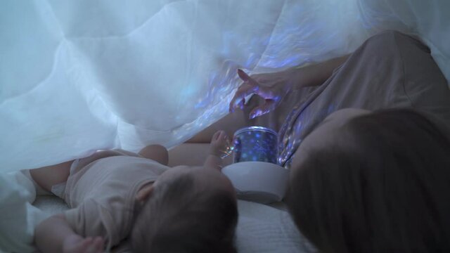 asian woman mother parent playing starry sky night light projector with baby infant under blanket in bedroom at home. pointing star teaching baby a lesson before bed time.