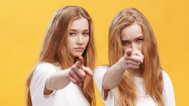 Its your fault. Two serious redhaired twin sisters pointing fingers to camera, blaming you, orange studio background