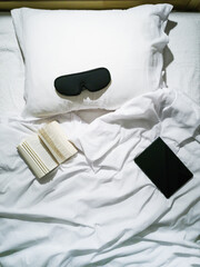 On the bed lies a tablet, an open book, and a sleep mask