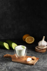 Obraz na płótnie Canvas Greek tzatziki sauce in a glass glass in a double bottom on a wooden rustic board with whole garlic, cucumber and lemon on a dark background