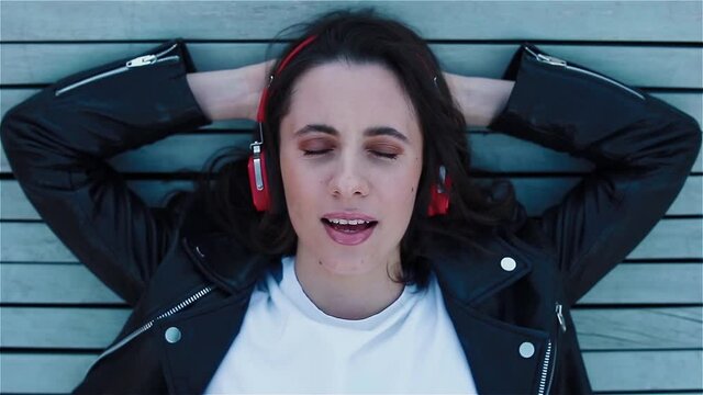 Young smiling woman in black leather jacket listening to music in big red headphones lying on the bench and singing a song ooking to the camera. Top view..