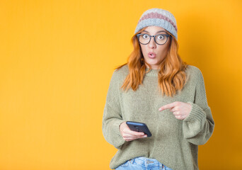 Surprised young woman shows to her phone unexpected incredible notification, isolated on yellow background. Amazed girl. 5g concept