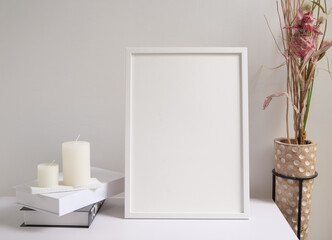 Mock up poster frame black and white books dried Lagurus ovatus flowers composition in modern glass vase and candles on  white table and cement wall background