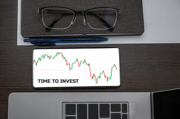 Buy or sell stocks concept. Top view of stocks price candlestick chart in phone on table near laptop, notepad and glasses with inscription time to invest. Business, finance concept.