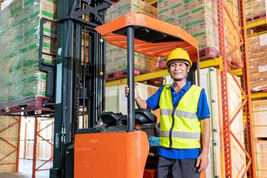 Forklift driver asian elderly man in safety jumpsuit uniform with yellow hardhat at warehouse. Worker male senior looking at camera and smiling in forklift loader work