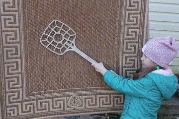 A girl preschooler knocks out a small carpet with a flapper in the street. Home help