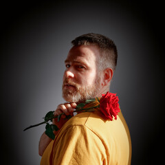 An attractive bearded calm man in a yellow T-shirt holds a red rose on his shoulder and looks thoughtfully aside. The face is illuminated by the light from the window.