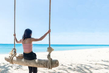 Back Beautiful young Asian women with tan skin wearing pink tank black pants sitting on timber swing. beach and seaside fresh sky background. Holiday, Vacation, Chilling, Relax concept. Alone Concept.