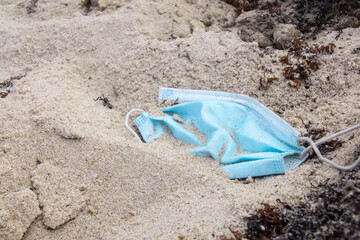 A discarded pale blue surgical medical face mask lying in the sand on a beach in Barbados, surrounded by seaweed. Partially buried. Concept - don't litter, plastic pollution, ocean clean up, COVID-19.