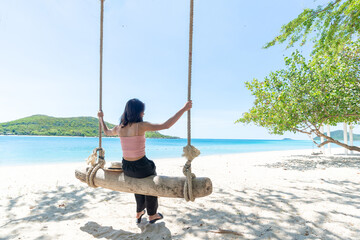 Back Beautiful young Asian women with tan skin wearing pink tank black pants sitting on timber swing. beach and seaside fresh sky background. Holiday, Vacation, Chilling, Relax concept. Alone Concept.