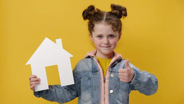 Beautiful little preschool girl holding small paper white house model with thumb up like gesture, wears denim jacket, isolated over yellow background wall with copy space. Housing and mortgage concept