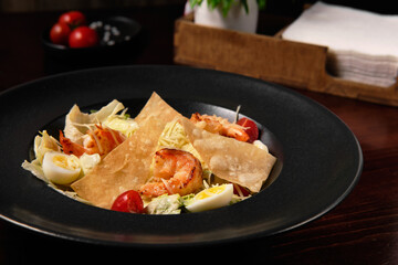 Appetizing Caesar salad with fried shrimps in a black plate on a dark wooden background. In the background there is a beautiful presentation and table setting. Healthy and dietary food.