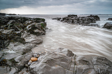Long exposure of waves streaming through rock formations near the shoreline.  There is an orange coloured pebble, noticeable against the grey rocks